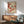 May include: Japanese-style artwork showing a small bird with black cap and wings perched on branches of red berries, displayed in thin wooden frame above mid-century modern credenza with table lamp, next to sage green upholstered armchair with wooden arms in sunlit room with white walls