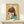 May include: Framed artwork displaying a stylized illustration of a young girl with brown bob haircut and large yellow-green eyes, created using mixed media with collaged document fragments on yellow background. Wooden frame sits on white wooden surface beside small wrapped gift with evergreen sprig. Minimalist white wall background.