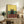 May include: Framed poster print featuring a black cat with yellow eyes sitting on a wooden surface between two wine glasses and a wine bottle, with yellow and teal background colors. Displayed on rustic wooden sideboard with books, ceramic vase, and fern plant in modern living room setting with beige couch.
