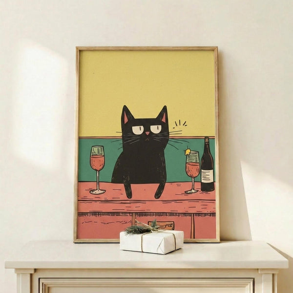 May include: Framed poster print displaying a black cat with half-closed eyes sitting on a wooden bar counter next to wine glasses filled with red wine and a dark bottle. The illustration features a yellow upper background, teal green wall, and coral pink wooden counter with drawers below. The artwork is shown in a thin wooden frame on a light wood surface alongside a small wrapped gift with evergreen sprig, against a white wall.