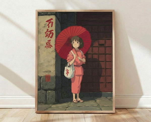 May include: A framed poster print in a light wood frame leaning against a white wall on light wooden flooring. The artwork shows an anime-style young girl with short brown hair wearing a pink outfit, holding a red traditional umbrella and white bag. She stands in front of a traditional Japanese building with red wooden doors and stone walls. Red Japanese characters appear on the left side of the image. The scene has warm, muted colors in a hand-drawn animation style.