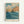 Vintage Japanese woodblock print style artwork showing dramatic blue ocean waves crashing against orange and tan colored rocky cliffs. A turquoise sky filled with white and gray clouds stretches across the top. The print features bold colors and traditional Japanese artistic techniques. Text at bottom reads "HASUI KAWASE" with Japanese characters and "The National Museum of Modern Art, Tokyo." The artwork is displayed in a light wooden frame against a white wall.