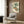 May include: Vintage Japanese botanical artwork showing persimmon fruit hanging from leafy branches, displayed in light wood frame on warm brown wood paneled wall. Modern beige upholstered chair with geometric patterned pillow sits below artwork, beside brass floor lamp and dark round side table with open book. Soft natural lighting from nearby window.