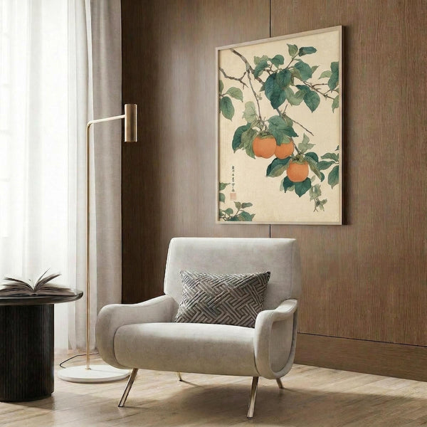 May include: Vintage Japanese botanical artwork showing persimmon fruit hanging from leafy branches, displayed in light wood frame on warm brown wood paneled wall. Modern beige upholstered chair with geometric patterned pillow sits below artwork, beside brass floor lamp and dark round side table with open book. Soft natural lighting from nearby window.
