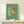 May include: Framed cartoon poster showing golden retriever wearing sunglasses sitting on green toilet reading pink newspaper in tiled bathroom. Light wood frame displayed on white shelf next to small wrapped gift with twine bow against cream wall.