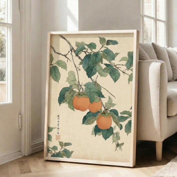 May include: Framed botanical artwork leaning against white wall next to beige sofa, showing orange persimmons hanging from green leafy branches in traditional Japanese watercolor style on cream background, with Asian calligraphy and red seal stamp in lower left corner, displayed in light wood frame on hardwood floors near large windows