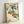 May include: Japanese botanical art print in light wood frame leaning against white wall, showing orange persimmons hanging from branches with detailed green leaves on cream background, traditional calligraphy and red seal stamp visible, natural lighting creating soft shadows