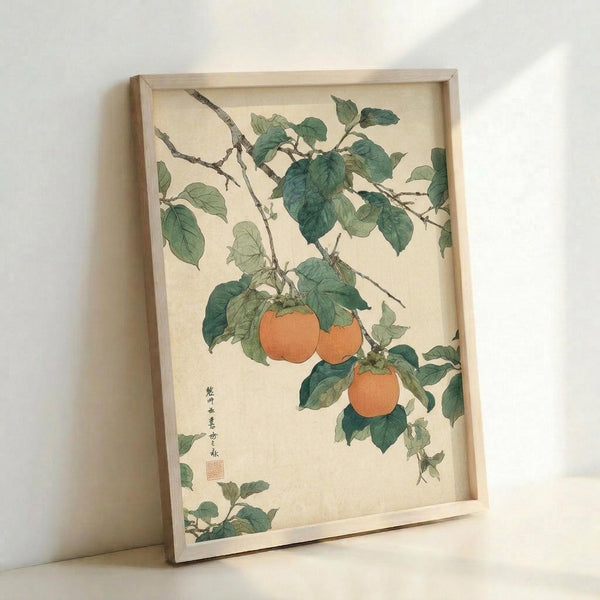 May include: Japanese botanical art print in light wood frame leaning against white wall, showing orange persimmons hanging from branches with detailed green leaves on cream background, traditional calligraphy and red seal stamp visible, natural lighting creating soft shadows