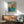 May include: A framed vintage Japanese woodblock print showing blue ocean waves, rocky coastal cliffs in warm orange and brown tones, and a cloudy turquoise sky. The artwork hangs above a mid-century modern wooden sideboard with sliding doors. A sage green upholstered armchair with wooden frame sits nearby, along with a white ceramic table lamp and small round coffee table. Natural sunlight creates geometric shadows on the white wall.