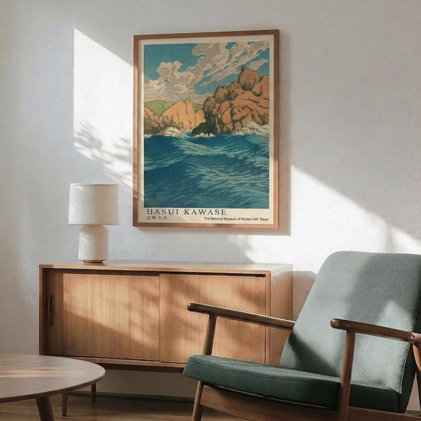 May include: A framed vintage Japanese woodblock print showing blue ocean waves, rocky coastal cliffs in warm orange and brown tones, and a cloudy turquoise sky. The artwork hangs above a mid-century modern wooden sideboard with sliding doors. A sage green upholstered armchair with wooden frame sits nearby, along with a white ceramic table lamp and small round coffee table. Natural sunlight creates geometric shadows on the white wall.