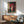 May include: Framed anime-style poster showing young girl in red kimono holding red parasol and white bag, standing by traditional Japanese building with wooden doors and stone walls. Displayed above wooden sideboard with table lamp in modern living room with gray upholstered chair and round coffee table.