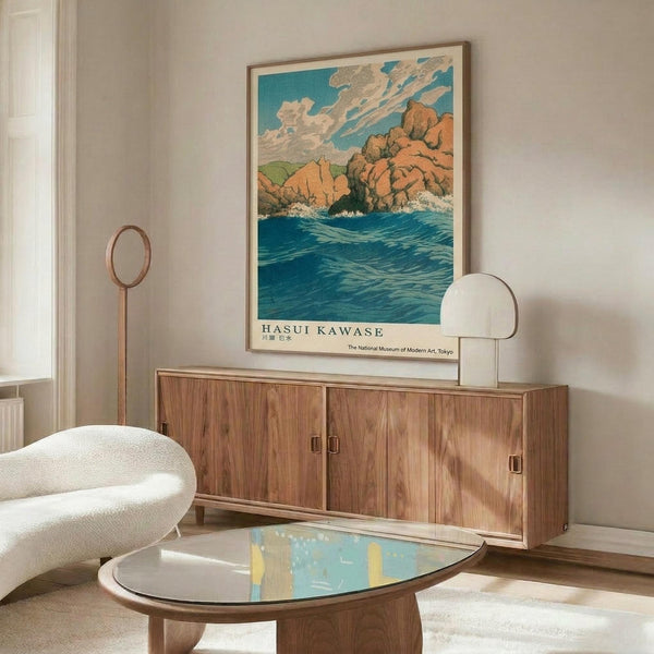 May include: Japanese woodblock print poster featuring turquoise ocean waves, orange rocky cliffs, and cloudy blue sky, displayed in thin wood frame above mid-century walnut credenza with sliding doors. White curved lounge chair and oval glass coffee table with brass legs visible in foreground. Warm natural lighting creates soft shadows on cream walls.
