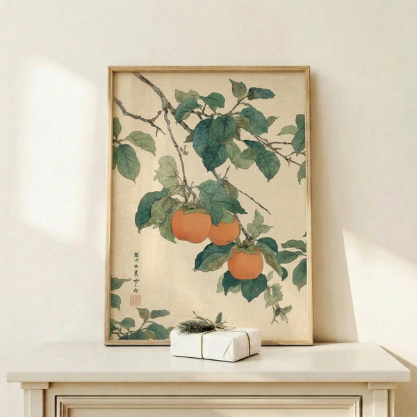 May include: Vintage Japanese botanical art print in thin gold frame showing orange persimmons hanging from green leafy branches with calligraphy, displayed on light wood console table next to small white wrapped gift with natural twig decoration against cream wall