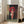 May include: Framed anime-style poster showing a young girl with brown bob haircut holding a red traditional umbrella, wearing coral pink clothing and carrying a white bag with red characters. She stands on stone pavement near a brick wall with yellow vertical banner containing red Japanese characters. The artwork is displayed in a light wooden frame leaning against a white wall next to a beige upholstered chair in a bright, naturally-lit room with white window frames.