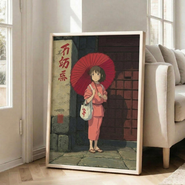 May include: Framed anime-style poster showing a young girl with brown bob haircut holding a red traditional umbrella, wearing coral pink clothing and carrying a white bag with red characters. She stands on stone pavement near a brick wall with yellow vertical banner containing red Japanese characters. The artwork is displayed in a light wooden frame leaning against a white wall next to a beige upholstered chair in a bright, naturally-lit room with white window frames.