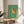 May include: Framed poster showing a golden retriever wearing sunglasses sitting on a toilet while reading a red book in a green-tiled bathroom. The cartoon-style artwork features bright colors with the dog on a white toilet against mint green wall and floor tiles, with a red wall-mounted dispenser. The wooden frame sits on a round wooden side table next to a beige upholstered chair with white pillow, plant leaves visible in corner.
