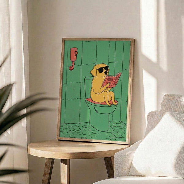 May include: Framed poster showing a golden retriever wearing sunglasses sitting on a toilet while reading a red book in a green-tiled bathroom. The cartoon-style artwork features bright colors with the dog on a white toilet against mint green wall and floor tiles, with a red wall-mounted dispenser. The wooden frame sits on a round wooden side table next to a beige upholstered chair with white pillow, plant leaves visible in corner.