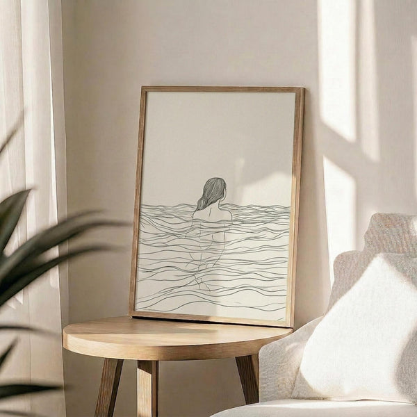 May include: Minimalist line drawing of woman submerged in wavy water, shown from behind with long hair, rendered in simple black lines on cream background. Displayed in thin wooden frame on round wooden side table next to beige upholstered chair. Soft natural lighting creates shadows on neutral wall with plant leaves visible in corner.