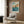 May include: A vintage Japanese woodblock print poster featuring rocky coastal cliffs, blue ocean waves, and cloudy sky in warm earth tones, displayed in a light wooden frame on wood-paneled wall. Modern reading nook with cream upholstered armchair, geometric patterned pillow, brass floor lamp, and dark round side table with open book creates cozy interior styling scene.