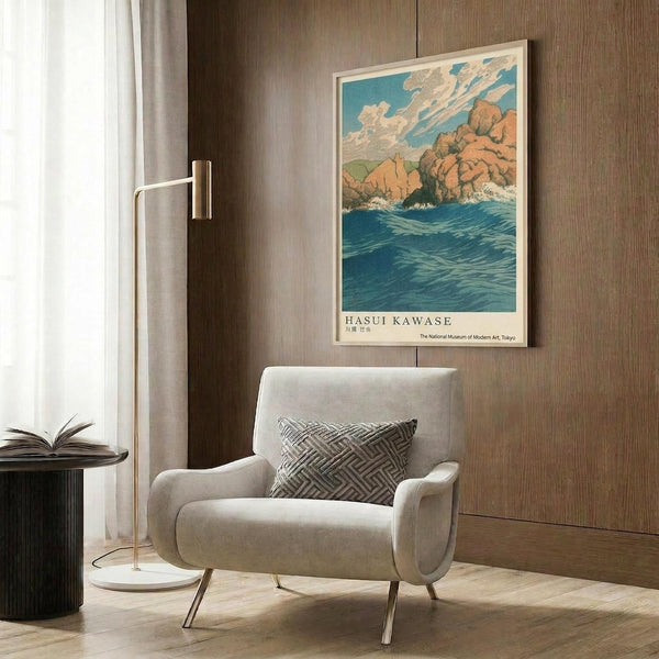 May include: A vintage Japanese woodblock print poster featuring rocky coastal cliffs, blue ocean waves, and cloudy sky in warm earth tones, displayed in a light wooden frame on wood-paneled wall. Modern reading nook with cream upholstered armchair, geometric patterned pillow, brass floor lamp, and dark round side table with open book creates cozy interior styling scene.