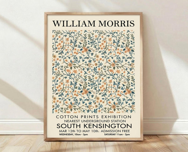 May include: Framed poster with William Morris floral pattern in peach and blue tones, featuring intricate botanical design with curving vines and blooms. Exhibition details for Cotton Prints show in South Kensington displayed below artwork. Light wood frame sits on wooden floor against white textured wall with soft natural lighting casting diagonal shadows.