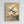 May include: Framed botanical art print hanging on white wall showing magnolia branch with pale cream and pink blossoms against warm beige background. Traditional Asian-style painting with delicate brushwork, dark twisted branches, and green leaves. Natural wood frame. Japanese calligraphy and red seal stamps in upper right corner.