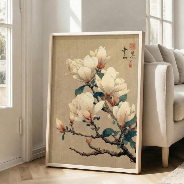 May include: Large framed botanical art print leaning against white wall next to beige upholstered chair. Artwork features delicate white magnolia blossoms with pink-tinted edges on dark branches with green leaves, painted in traditional Japanese style on textured beige background. Light wooden frame, natural lighting from nearby windows, hardwood floors visible.