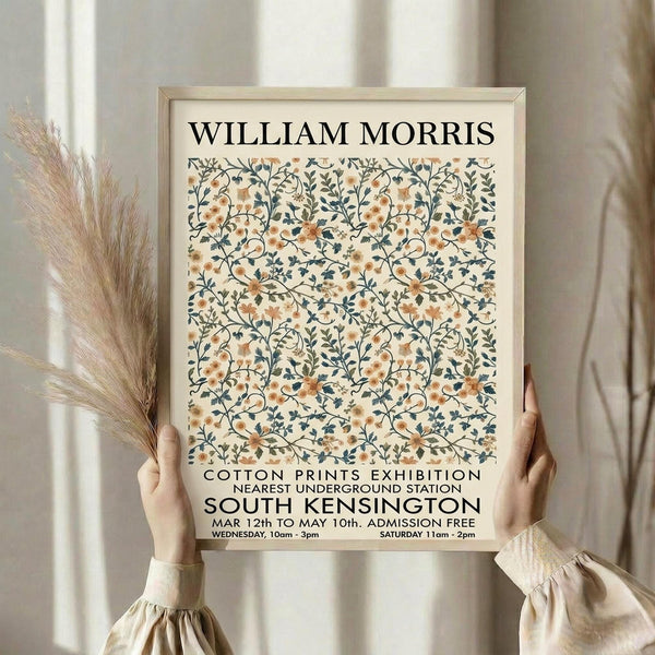 May include: Person in cream blouse holding framed poster featuring William Morris-style floral pattern in peach and navy blue tones. Light wood frame against neutral background with dried pampas grass visible. Exhibition poster shows intricate botanical design with curved stems, small flowers, and leaves in vintage Arts and Crafts style.