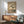May include: Japanese-style botanical artwork showing white magnolia blossoms on branches against a tan background, displayed in a thin wooden frame above a mid-century modern credenza. The scene features a sage green upholstered armchair with wooden arms, a small round side table, and a white ceramic table lamp. Natural sunlight creates dramatic shadows across the white wall, highlighting the artwork's delicate floral details and traditional calligraphy in the upper right corner.