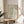 May include: Framed vintage-style exhibition poster with William Morris floral pattern in sage green, peach, and cream tones displayed on a round wooden side table next to a beige upholstered chair. Natural sunlight streams across white walls, with a partial view of a potted plant. The poster features intricate botanical designs and exhibition text for South Kensington Cotton Prints show.