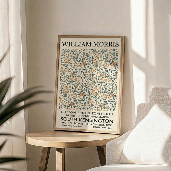 May include: Framed vintage-style exhibition poster with William Morris floral pattern in sage green, peach, and cream tones displayed on a round wooden side table next to a beige upholstered chair. Natural sunlight streams across white walls, with a partial view of a potted plant. The poster features intricate botanical designs and exhibition text for South Kensington Cotton Prints show.