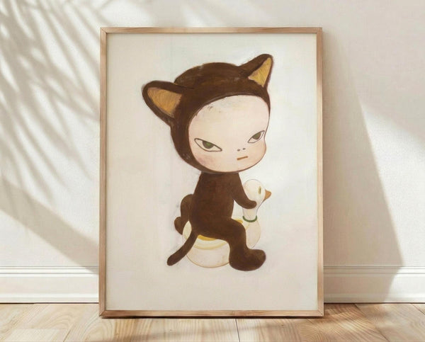 May include: Framed art print displaying a stylized character in a brown cat costume with pointed ears and tail, sitting and holding what appears to be a small white duck. The illustration features soft, muted colors with the character having a pale face and contemplative expression. The artwork is shown in a light wood frame against a cream background, positioned on wooden flooring with natural sunlight casting diagonal shadows across the white wall.