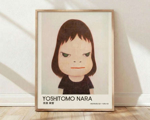 May include: Framed poster print leaning against white wall on light wood floor. Features minimalist illustration of young girl with brown bob haircut, green eyes, and serious expression wearing dark clothing. Clean geometric art style with muted colors on cream background. Text shows "YOSHITOMO NARA" and Japanese characters with exhibition dates. Natural wood frame, soft lighting creates diagonal shadows on textured white wall.