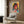 May include: Framed cyberpunk-style portrait poster displayed on wood paneled wall above beige armchair with geometric pillow. Artwork features woman with pink-tinted sunglasses in vibrant yellow, pink, and teal colors with digital glitch effects and Japanese text elements. Modern reading nook setting with brass floor lamp and side table.