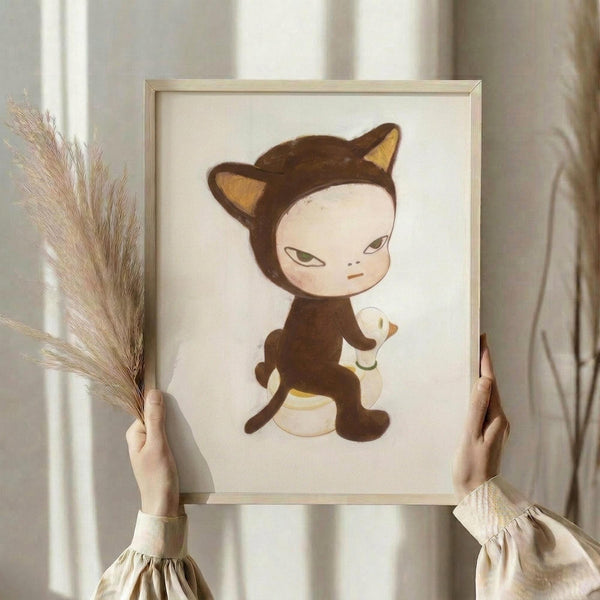 May include: Hands holding a framed art print featuring a cute character in a brown cat costume with pointed ears and tail, riding a white duck. The illustration has a soft, whimsical style with muted colors on cream paper. Person wears a beige ruffled sleeve top and holds dried pampas grass in one hand. Natural lighting creates gentle shadows on white curtained background.