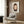 May include: Framed poster featuring a stylized portrait of a young girl with brown bob haircut and green eyes in minimalist art style, displayed above a beige upholstered armchair with geometric patterned pillow. Modern living room setting with wood paneled walls, brass floor lamp, and side table with open book. Warm neutral tones throughout the space with natural lighting from window.