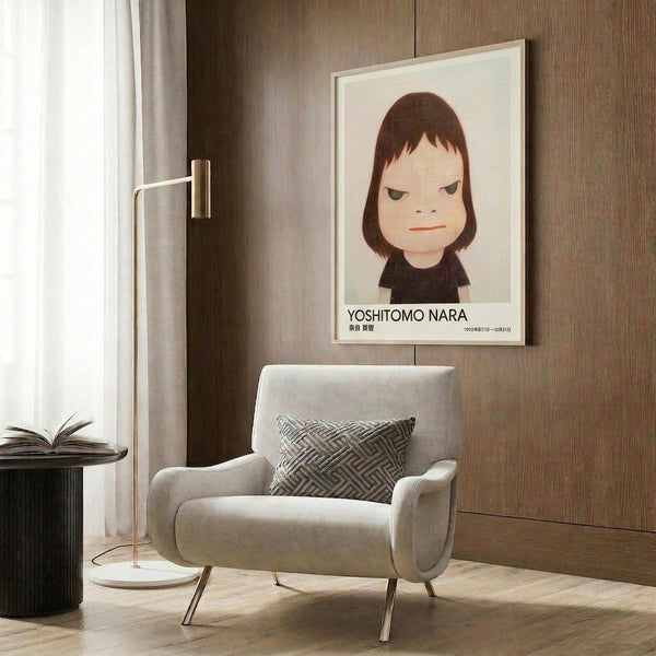 May include: Framed poster featuring a stylized portrait of a young girl with brown bob haircut and green eyes in minimalist art style, displayed above a beige upholstered armchair with geometric patterned pillow. Modern living room setting with wood paneled walls, brass floor lamp, and side table with open book. Warm neutral tones throughout the space with natural lighting from window.