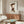 May include: Framed poster featuring a stylized cartoon character in a brown cat costume with pointed ears and yellow accents, displayed above a mid-century modern wooden sideboard. The artwork shows a pale-faced figure in a minimalist art style against a white background. Room setting includes a white curved chair, oval glass coffee table with colorful abstract design, white mushroom-shaped table lamp, and neutral beige walls with natural lighting.
