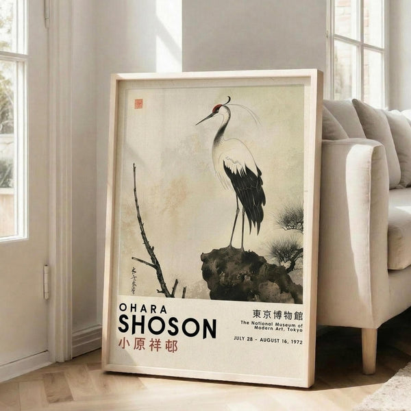 May include: Framed vintage Japanese art poster featuring a white crane with black wing tips and red head detail standing on a dark rock beside bare branches, displayed against a light wooden frame in a bright living room with beige sofa, white walls, and large windows casting natural light across hardwood floors.