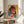 May include: Framed cyberpunk-style artwork featuring a woman with round glasses and dark hair against vibrant yellow, pink, and blue geometric backgrounds with Japanese text elements. The poster sits on a light wood round side table next to a beige upholstered chair with white throw pillow. Natural sunlight creates shadows across white walls, with green plant leaves visible in the corner.