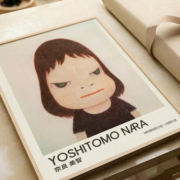 May include: A framed art print featuring a stylized illustration of a young girl with shoulder-length brown hair, green eyes, and a subtle frown, rendered in muted tones with a minimalist aesthetic. The print has exhibition-style text at the bottom reading "YOSHITOMO NARA" with Japanese characters and dates from 1992. The artwork is displayed in a light wooden frame on a cream-colored surface next to white fabric or ribbon.