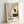 May include: Framed vintage exhibition poster featuring a Japanese crane painting in traditional ink wash style, showing an elegant white and black crane with red head crest perched on dark rocks beside bare branches. The cream-colored poster displays museum text and Japanese characters below the artwork. Light wood frame leans against white wall with soft natural lighting creating gentle shadows.