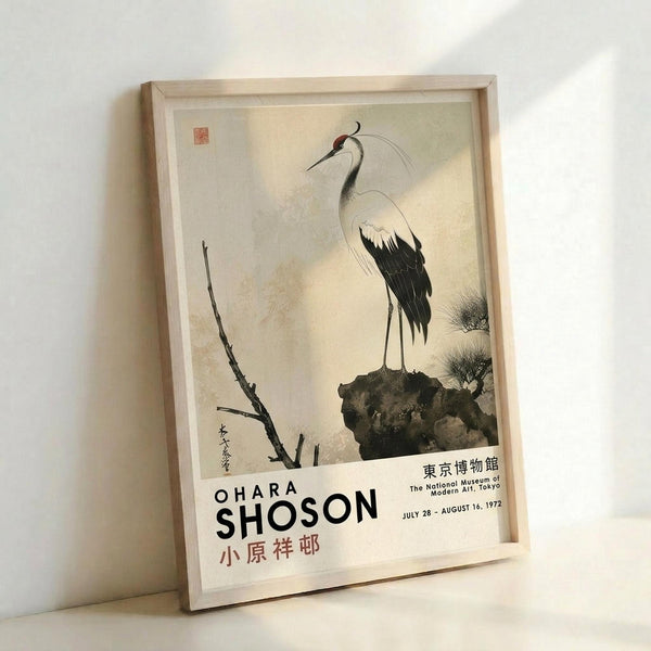 May include: Framed vintage exhibition poster featuring a Japanese crane painting in traditional ink wash style, showing an elegant white and black crane with red head crest perched on dark rocks beside bare branches. The cream-colored poster displays museum text and Japanese characters below the artwork. Light wood frame leans against white wall with soft natural lighting creating gentle shadows.