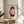 May include: Framed poster featuring a minimalist illustration of a young girl with brown bob haircut, green eyes, and orange lips wearing a dark shirt, displayed on a round wooden stool next to a beige upholstered chair and potted plant in a bright room with natural lighting.