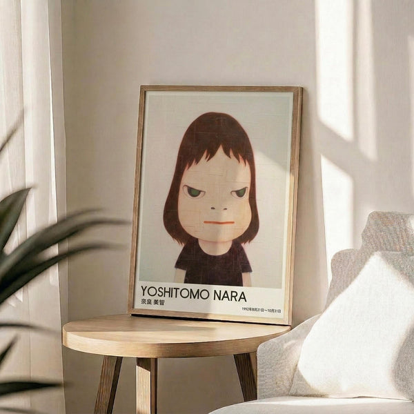 May include: Framed poster featuring a minimalist illustration of a young girl with brown bob haircut, green eyes, and orange lips wearing a dark shirt, displayed on a round wooden stool next to a beige upholstered chair and potted plant in a bright room with natural lighting.