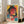 May include: Framed cyberpunk-style poster featuring a woman with round glasses and dark hair in vibrant yellow, pink, and teal colors with Japanese text overlay. The artwork has a distressed, layered collage aesthetic with geometric elements. White wooden frame leans against a bright living room wall next to a beige sectional sofa with windows showing natural light.