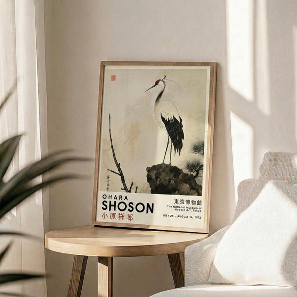 May include: Framed vintage Japanese art print featuring a white and black crane perched on a rock beside bare branches, displayed on a round wooden side table next to a beige upholstered chair. Soft natural lighting creates geometric shadows on the white wall behind. A plant's leaves are visible in the foreground, adding depth to this minimalist modern interior setting.
