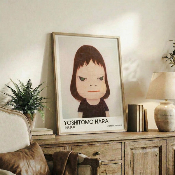 May include: Framed art print featuring a stylized illustration of a young girl with brown bob haircut and green eyes, wearing a dark shirt against a light background. The poster displays "YOSHITOMO NARA" text at bottom. Shown in thin wooden frame, positioned on rustic wooden sideboard alongside fern plant, ceramic vase, and books in neutral-toned living room setting.