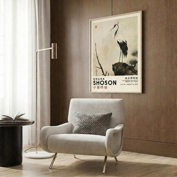 May include: Framed vintage Japanese crane artwork hanging on wood-paneled wall above beige upholstered armchair with geometric pillow. Traditional ink painting shows white crane with black wing tips standing on dark rock formation. Exhibition poster format with "OHARA SHOSON" text below artwork. Modern reading nook setting with brass floor lamp, small side table with open book, and neutral curtains.