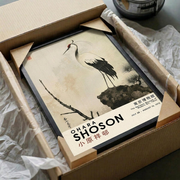 May include: Japanese crane art poster in black frame being unpacked from brown cardboard box with white tissue paper. The vintage-style print shows a white crane with black wing details standing on dark ground against cream background. Wooden dowels visible alongside the framed artwork on white fabric surface.